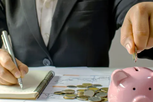 Pessoa de fato a escrever num caderno enquanto coloca uma moeda num mealheiro rosa, com moedas e gráficos sobre a mesa